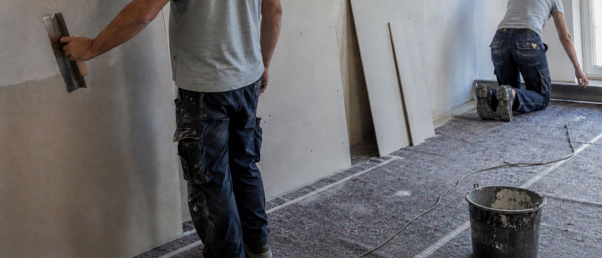 Zwei Personen bei Ausbauarbeiten in einem Altbauraum, von hinten aufgenommen: links wird eine Wandfläche gespachtelt, rechts eine Bodenfläche kontrolliert